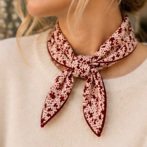 May include: A square neck scarf featuring a dark red and cream-colored intricate geometric pattern. The scarf is tied in a simple knot at the center, displaying a repeating star-like motif across the fabric with a solid dark red border edge.
