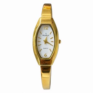 May include: A gold-tone quartz wristwatch featuring an elongated oval case and a white dial. The face displays the brand name Peugeot, the word Quartz, and gold-colored hour markers with the numbers 12 and 6 in a gold-tone font style.