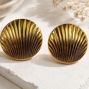 May include: A pair of circular gold-toned earrings featuring a raised, radiating fan-like shell pattern. The surface of each earring is textured with fine, vertical ridges that extend from the center to the outer edge, creating a metallic sheen.