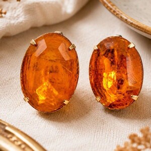 May include: A pair of oval-shaped earrings featuring faceted, translucent orange stones. Each stone is secured by four small gold-toned metal prongs that hold the gem in place, creating a classic and symmetrical design for these jewelry pieces.
