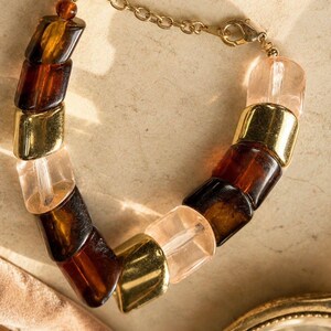 May include: A bracelet featuring alternating chunky beads in translucent amber, clear pale pink, and polished gold-tone metal. The beads are strung together and finished with a gold-tone metal chain and a lobster claw clasp closure.