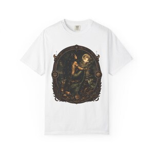 May include: A white short-sleeve t-shirt featuring a central graphic print of a mermaid sitting on a rock under a full moon. The illustration is enclosed in an ornate, dark-toned oval frame with small trident and seashell icons on the sides.