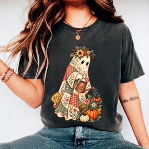 Puede incluir: Una camiseta gris carbón con el gráfico de un fantasma envuelto en una colcha de retazos con patrones rojos, blancos y verdes. El fantasma lleva una corona floral y usa una regadera marrón para regar un grupo de flores coloridas y calabazas.