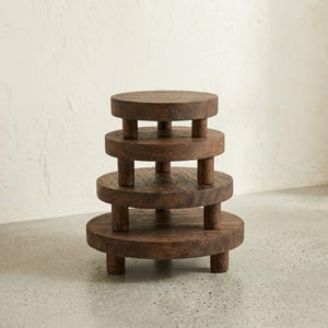 May include: A dark brown, tiered wooden display stand with four circular platforms of decreasing size. Each platform is supported by short, cylindrical legs, creating a multi-level structure. The wood grain is visible, adding texture to the piece.