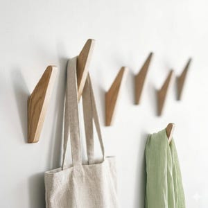 Pode incluir: Um conjunto de ganchos de parede de madeira com um design em forma de seta. Os ganchos são montados numa parede branca, com uma sacola bege e um lenço verde claro pendurados em alguns deles. Os ganchos são feitos de madeira clara.