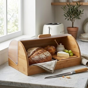 Puede incluir: Una panera de bambú con tapa transparente y curvada, que contiene un pan, un muffin, queso y uvas. También hay una tabla de cortar de madera y un cuchillo de pan. La panera está sobre una encimera de mármol.