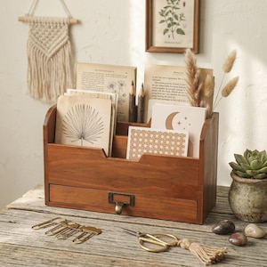 Puede incluir: Un organizador de escritorio de madera con múltiples compartimentos y un cajón. El organizador está lleno de tarjetas de estilo vintage, lápices y plantas secas. Clips dorados y tijeras están sobre la superficie de madera frente al organizador. Un pequeño tapiz de macramé está en el fondo.