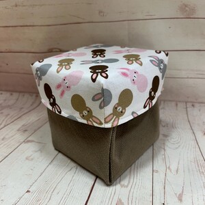 May include: A square storage box with a brown canvas exterior and a white fabric top featuring a repeating pattern of brown, pink, and grey bunnies.