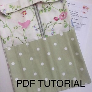 Peut inclure: Une trousse à aiguilles à tricoter en tissu avec un motif floral et un fond à pois verts. Le rabat supérieur présente un oiseau rose et un motif floral. Le texte "PDF TUTORIAL" est imprimé sur le devant. Deux aiguilles à tricoter sont visibles à l'intérieur.