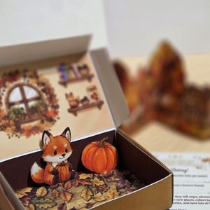 Può includere: Una piccola scatola di cartone contenente una sagoma di carta di una volpe che tiene una zucca, una sagoma di zucca separata e una base stampata con foglie autunnali. Il coperchio interno presenta un'illustrazione di una finestra e scaffali.