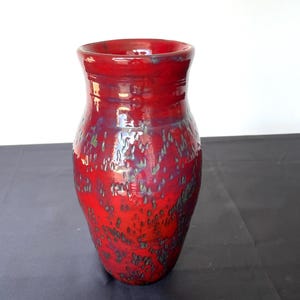 May include: A handmade red ceramic vase with a glossy finish. The vase has a wide base that tapers upwards, with a slightly flared rim. The surface is textured with dark green and blue accents, adding depth and visual interest to the piece.