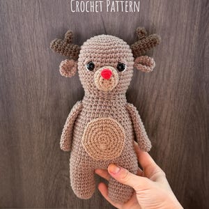 May include: A crocheted reindeer doll with brown yarn, featuring a red nose, black eyes, and small antlers. The doll has a circular design on its belly. Text on the image reads "RIAN THE REINDEER CROCHET PATTERN" and "CROCHET CRITTER CLUB."