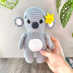 Pode incluir: Um coala de crochê cinza feito à mão com uma barriga branca e uma flor amarela atrás da orelha. O coala tem olhos pretos e nariz e boca rosa. O coala é um bicho de pelúcia.