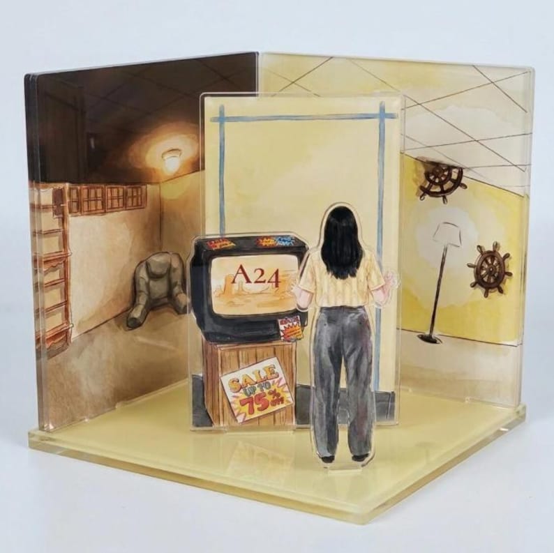 May include: A multi-layered acrylic diorama featuring a person with dark hair standing before a vintage television set. The screen displays the text A24, and a sign on the wooden stand reads SALE UP TO 75%. The background depicts a room with yellow walls.