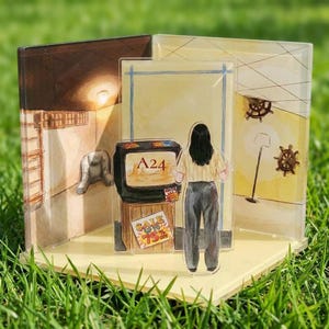 Puede incluir: Un diorama acrílico multicapa que muestra a una persona de pie frente a un televisor antiguo. La pantalla muestra el texto A24, mientras que un cartel debajo dice SALE UP TO 75% OFF. Los paneles de fondo representan una habitación con una lámpara.