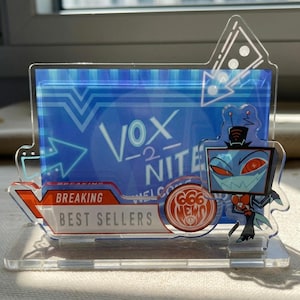 Könnte beinhalten: Transparenter Acrylständer mit blauem Hintergrund und dem Text "Vox -2- Nite". Enthält eine Cartoonfigur, die Worte "Breaking Best Sellers" und das Logo "666 News".