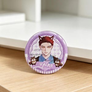May include: A circular pinback button featuring an anime-style illustration of a person with reddish-brown hair and cat ears. The background is light purple with a checkered pattern. The name VALKO is printed in white text at the bottom.