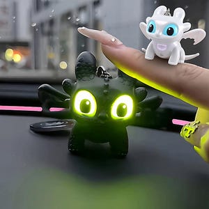 May include: Two dragon figurines, one black and one white, with glowing eyes. The black dragon is on a surface, while the white dragon is on a finger. Both have wings and are keychains. The black dragon's eyes glow green, and the white dragon's eyes glow blue.
