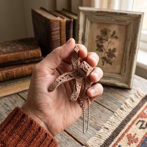 May include: A brown, detailed keychain in the form of a figure, held in a hand. The keychain features an intricate, openwork design and is connected to a silver chain. The background includes antique books and a framed picture.