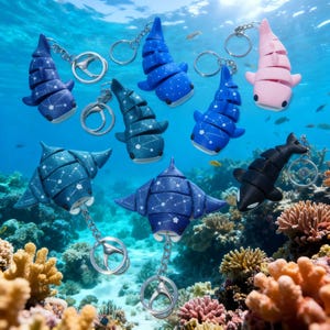 May include: Collection of marine animal keychains, including whale sharks, manta rays, and an orca. The keychains are in shades of blue, pink, and black, with silver key rings. The background shows an underwater coral reef scene.