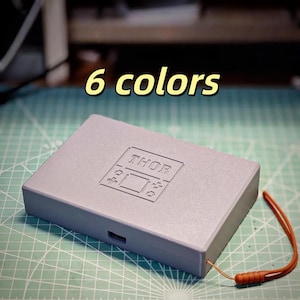 Könnte beinhalten: Eine rechteckige, hellgraue Box mit dem Wort "THOR" auf dem Deckel. Die Box hat ein kleines orangefarbenes Schlüsselband. Der Text "6 Farben" wird in Gelb über der Box angezeigt. Die Box befindet sich auf einer grünen Schneidematte.