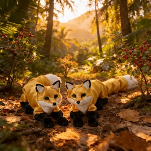 May include: Two plush fox toys with golden fur, white-tipped tails, and black paws are positioned in a forest. The foxes have black-lined ears and green eyes. The background features lush greenery and sunlight.