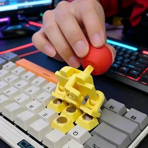 May include: A yellow mechanical keyboard keycap puller is shown with a red ball-shaped keycap. The keycap puller is designed to remove keycaps from a keyboard. The keyboard has white, gray, and orange keycaps.