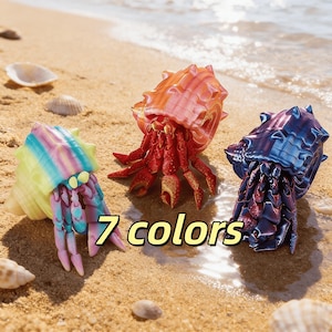 Puede incluir: Tres figuras de cangrejos ermitaños coloridos en una playa de arena. Uno es de color arcoíris, otro es rojo y naranja, y el tercero es azul y morado. El texto "7 colores" se muestra en fuente amarilla.
