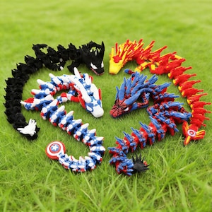 May include: A collection of colorful, articulated dragon toys arranged on green grass. The dragons feature various designs, including red, blue, and black, with some resembling comic book characters. Each dragon has a unique head and body design.