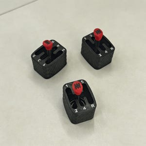 May include: Three black miniature gear shift knobs with red tops are arranged on a light surface. Each knob has white numbers and letters indicating gear positions. The gear shift knobs are likely 3D printed.