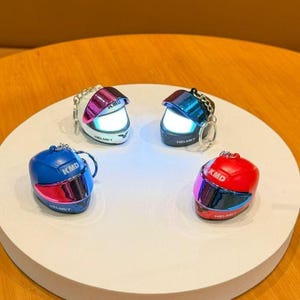 Op de afbeelding: Vier miniatuurhelm-sleutelhangers in wit, blauw, rood en zwart. Elke helm heeft een reflecterend vizier en de tekst "KMD" en "HELMET". De sleutelhangers zijn gerangschikt op een wit oppervlak.