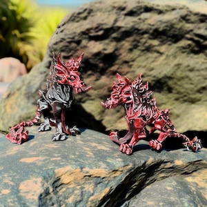 Puede incluir: Dos figuras detalladas de dragones, una negra y roja, la otra roja, posan sobre una roca. Los dragones tienen diseños intrincados, con rasgos afilados y una apariencia estilizada. Las figuras se colocan contra un fondo natural borroso.