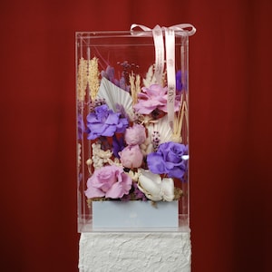 May include: A clear acrylic box filled with an arrangement of purple, pink, and white flowers. The flowers are arranged in a light blue box, and the arrangement is tied with a ribbon. The background is a solid red.