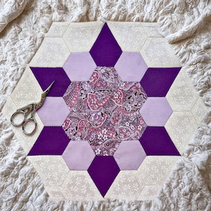 May include: A handmade, hexagonal quilt block with a central paisley pattern in pinks and purples. The design features purple, lavender, and cream-colored fabric pieces. A pair of gold-handled scissors rests on the quilt block.