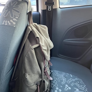 Könnte beinhalten: Ein grüner Canvas-Rucksack mit braunen Lederriemen hängt an der Kopfstütze eines Autositzes. Der Rucksack hat mehrere Taschen und einen Reißverschluss. Der Autositz ist grau mit einem weißen und grauen Muster.
