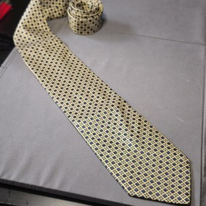 May include: A necktie featuring a repeating geometric pattern of small dark blue squares outlined in gold, set against a light gold background. The tie is laid out flat with the narrow end coiled at the top and the wide blade extending downward.