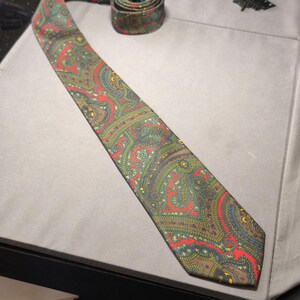 Può includere: Una cravatta con un dettagliato motivo paisley nei toni del verde oliva, rosso brillante e giallo. La cravatta è parzialmente arrotolata nella parte superiore, mostrando una consistenza liscia del tessuto e un'estremità a punta, con un righello nero.