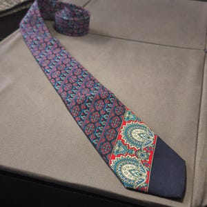 Può includere: Una cravatta fantasia con base blu navy e un motivo ripetuto in rosso, blu e verde acqua. La cravatta presenta un motivo paisley sulla punta. La cravatta è arrotolata nella parte superiore.