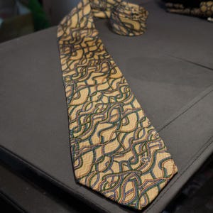 Puede incluir: Una corbata beige con un patrón repetido de líneas entrelazadas azules, verdes y moradas. La corbata está hecha de una tela suave y se muestra sobre una superficie gris.