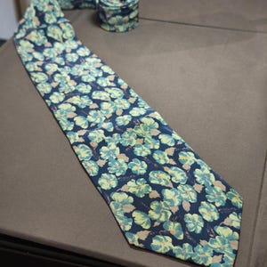 Può includere: Una cravatta blu navy con un motivo floreale ripetuto in azzurro e giallo pallido. La cravatta è parzialmente arrotolata e il motivo floreale è visibile anche sulla parte arrotolata.