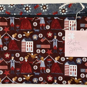 May include: A rectangular fabric pouch with a dark red background featuring a repeating pattern of colonial figures, American flags, eagles, and buildings. The top border is a blue floral print. A pink note with the text Leftie Zipper is attached.