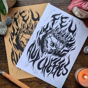 May include: Two rectangular art prints featuring a black ink block print design of a skull wearing a crown with an anarchy symbol, surrounded by stylized flames. The text "FEU AUX CHEFFES" is integrated into the design on both white and tan paper.
