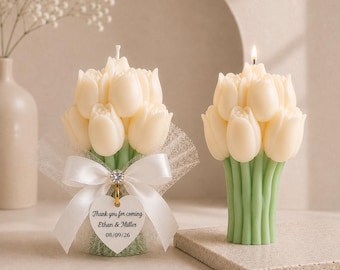 Op de afbeelding: Twee crèmekleurige tulpvormige kaarsen met groene stelen. Eén kaars is voorzien van een witte tule wikkel, een witte satijnen strik en een hartvormig label met de tekst: Thank you for coming, Ethan & Miller, 08/09/26. De andere kaars brandt.
