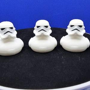 May include: Three white rubber duckies styled as Stormtroopers from Star Wars. Each duck has a white helmet with black facial features. They are arranged on a black circular surface against a blue background.