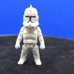May include: A small, white figurine of a Star Wars Clone Trooper. The figure is standing upright with a detailed helmet and armor. The background is a solid blue.