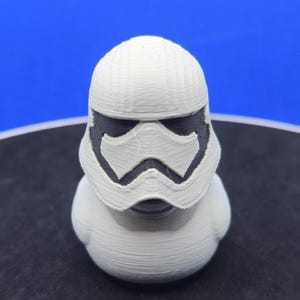 May include: A white, 3D-printed duck figurine with a Stormtrooper helmet design. The helmet features black accents around the eyes and mouth, and the duck has a smooth, rounded body. The background is a gradient of blue.