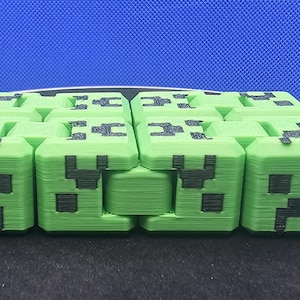 May include: A green, cube-shaped toy with a pixelated face design, resembling a character from a popular video game. The toy is made of multiple connected blocks, allowing for movement and manipulation. The design features black accents on a light green base.