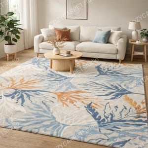 May include: A large rectangular area rug with a botanical design. The rug features a cream-colored background with a pattern of blue, orange, and white leaf-like shapes. The rug is placed in a living room setting with a white sofa and wooden coffee table.