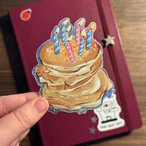 Könnte beinhalten: Ein holografischer Aufkleber mit einem Stapel Pfannkuchen mit brennenden blauen und rosa Kerzen. Der Aufkleber wird über einem weinroten Notizbuch mit einem sternförmigen Anhänger und anderen Aufklebern gehalten.