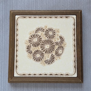 May include: Square decorative tile with a floral design in shades of brown and beige, set within a wooden frame. The tile features a central cluster of stylized flowers and a scalloped border.